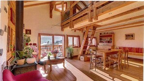 A spacious room with wooden beams, a fireplace, a dining area with a table and chairs, and a balcony with large windows.