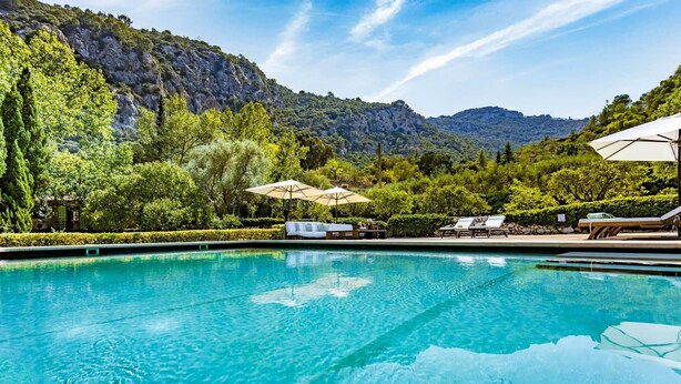 Una piscina cristallina con lettini e ombrelloni, circondata da vegetazione lussureggiante e montagne sullo sfondo.