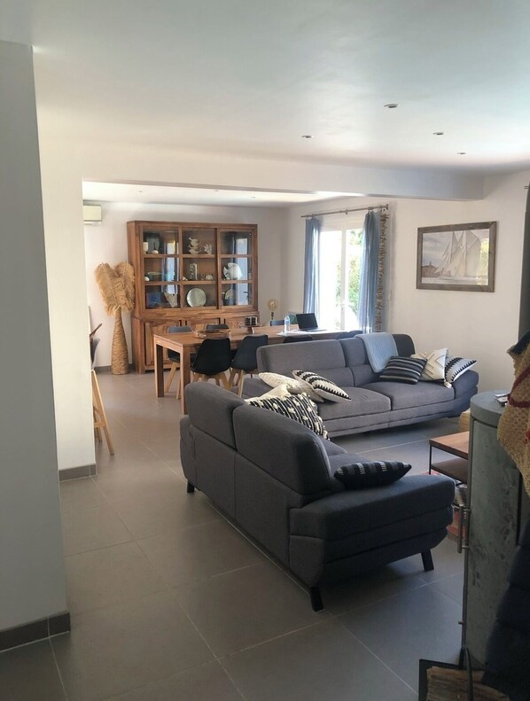 A spacious living room with a grey sofa, a wooden dining table with chairs, and a cabinet with glass doors.