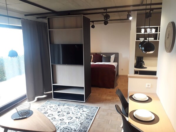 Ein kleines Zimmer mit einem Bett, einem Schreibtisch, einem Stuhl, einem Esstisch mit Stühlen, einem Bücherregal und einer Wandregal mit Küchenutensilien.