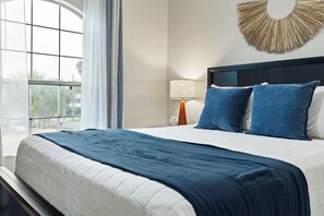 Et hotelværelse med en stor seng, et vindue med gardiner, en sengebordlampe og en dekorativ genstand på væggen.