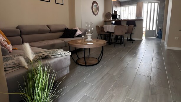 A modern living room with a sofa, a coffee table, and a potted plant. There are framed pictures on the wall and a kitchen area in the background.