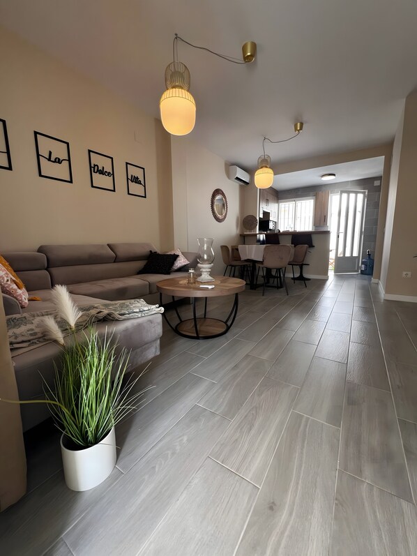 A modern living room with a sofa, a coffee table, and a potted plant. There are framed pictures on the wall and a kitchen area in the background.