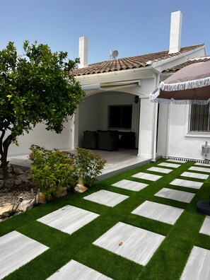 Uma casa com varanda coberta, jardim com grama sintética e pedras para caminhar, e vista para o quintal.