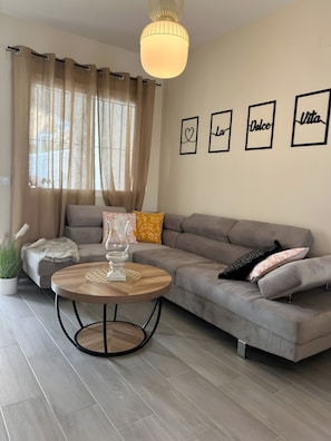 Uma sala de estar moderna com um sofá cinzento, uma mesa de centro redonda de madeira e uma luminária pendente.