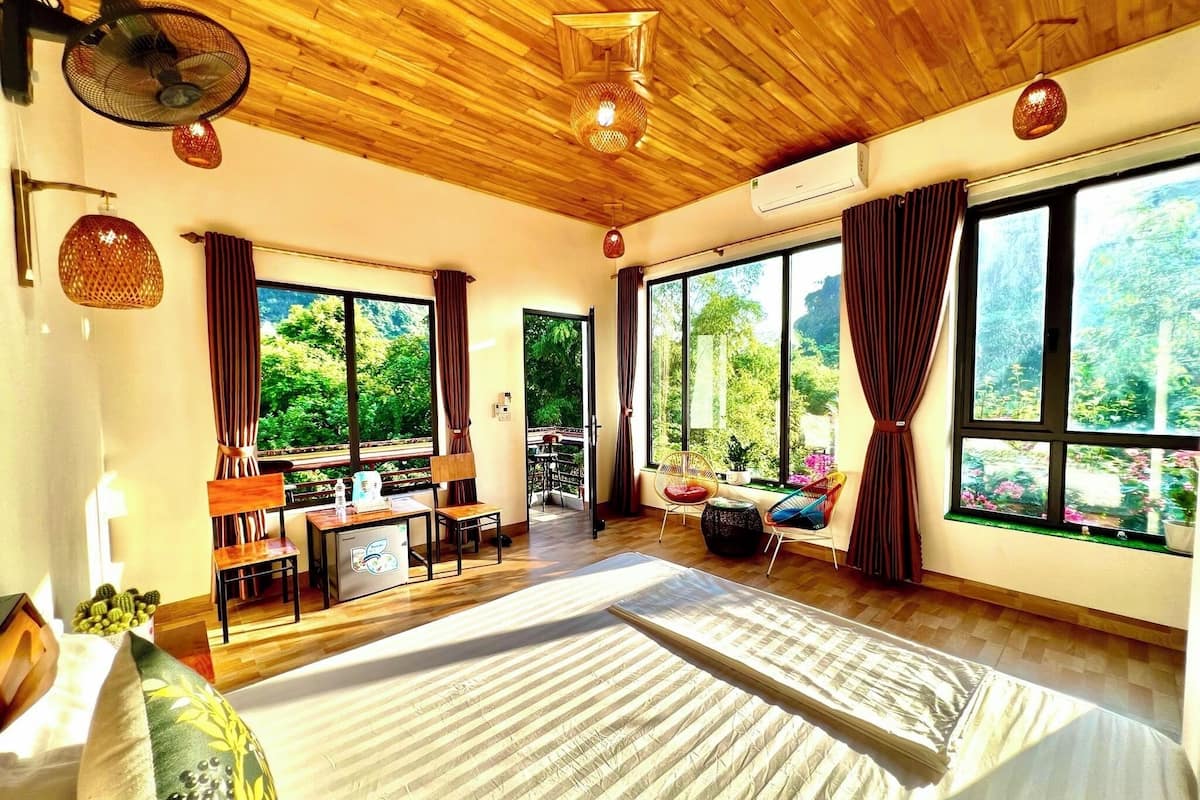 Une chambre dotée d’un grand lit, d’un bureau avec un ordinateur et offrant une vue sur la verdure grâce à de grandes fenêtres.