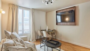 A modern living room with a sofa, a dining table with chairs, a flat-screen TV mounted on the wall, and a large window with curtains. - New apartment Center of Paris/Le Marais (Paris)