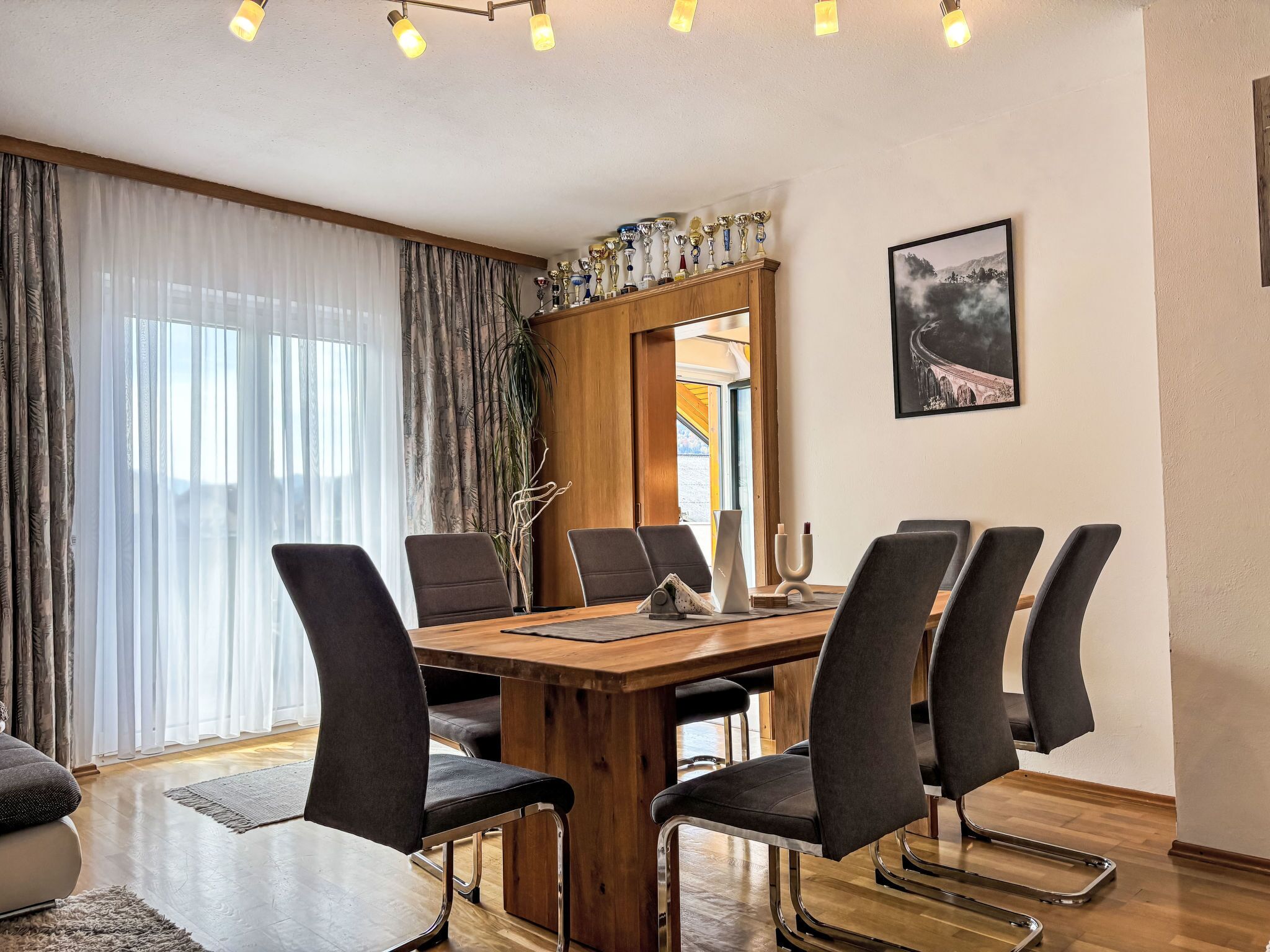 Ein Essbereich mit einem Holztisch und sechs Stühlen, einem großen Fenster mit Vorhängen sowie einem gerahmten Bild an der Wand.