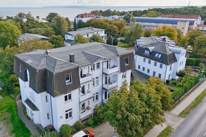 Kerrostalo, jossa on useita kerroksia, ja toisessa kerroksessa on parveke. Rakennuksen ympärillä on vehreää kasvillisuutta, ja parvekkeelta avautuu näkymä merelle.