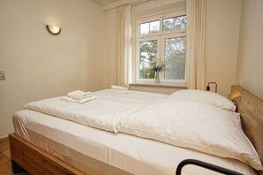 Ein Doppelbett mit weißer Bettwäsche, ein Nachttisch mit Lampe, ein Fenster mit Vorhängen und eine Wandleuchte.