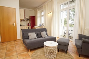 Ein modernes Wohnzimmer mit einer Couch, einem kleinen runden Tisch und einem Sessel. Im Hintergrund befindet sich eine Küchenzeile mit einem roten Schrank und einem Esstisch.