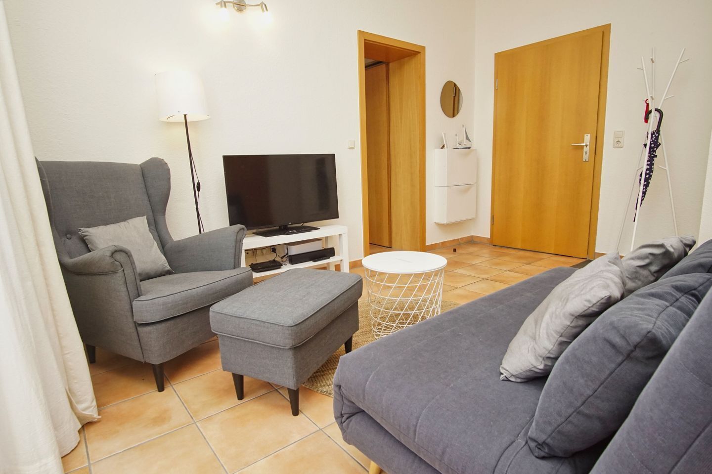A living room with a grey sofa, a grey armchair, a TV on a white stand, a small white round table, and a wooden door.