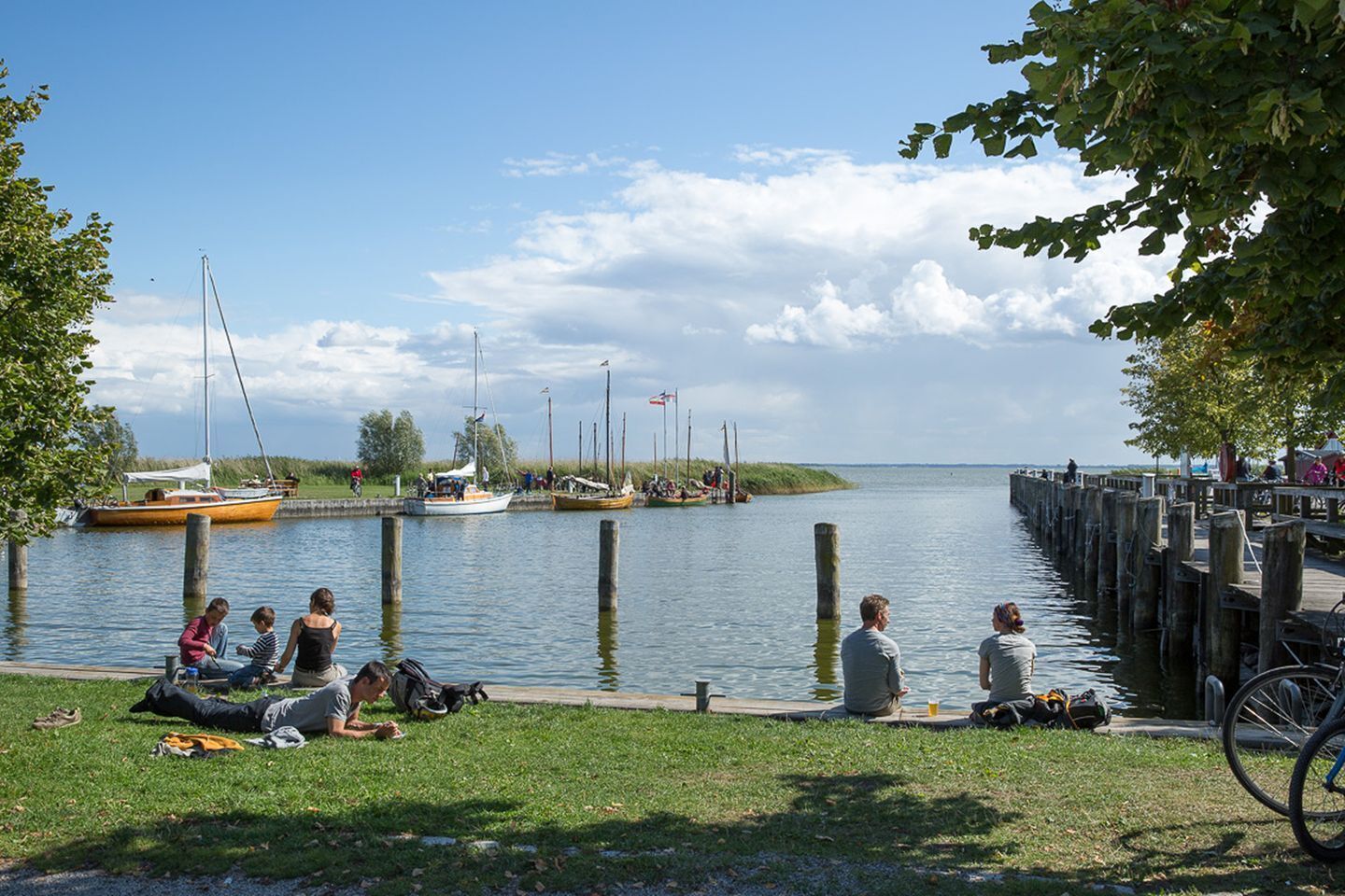 Escena junto al lago con barcos amarrados, gente relajándose sobre la hierba y un muelle de madera que se adentra en el agua.