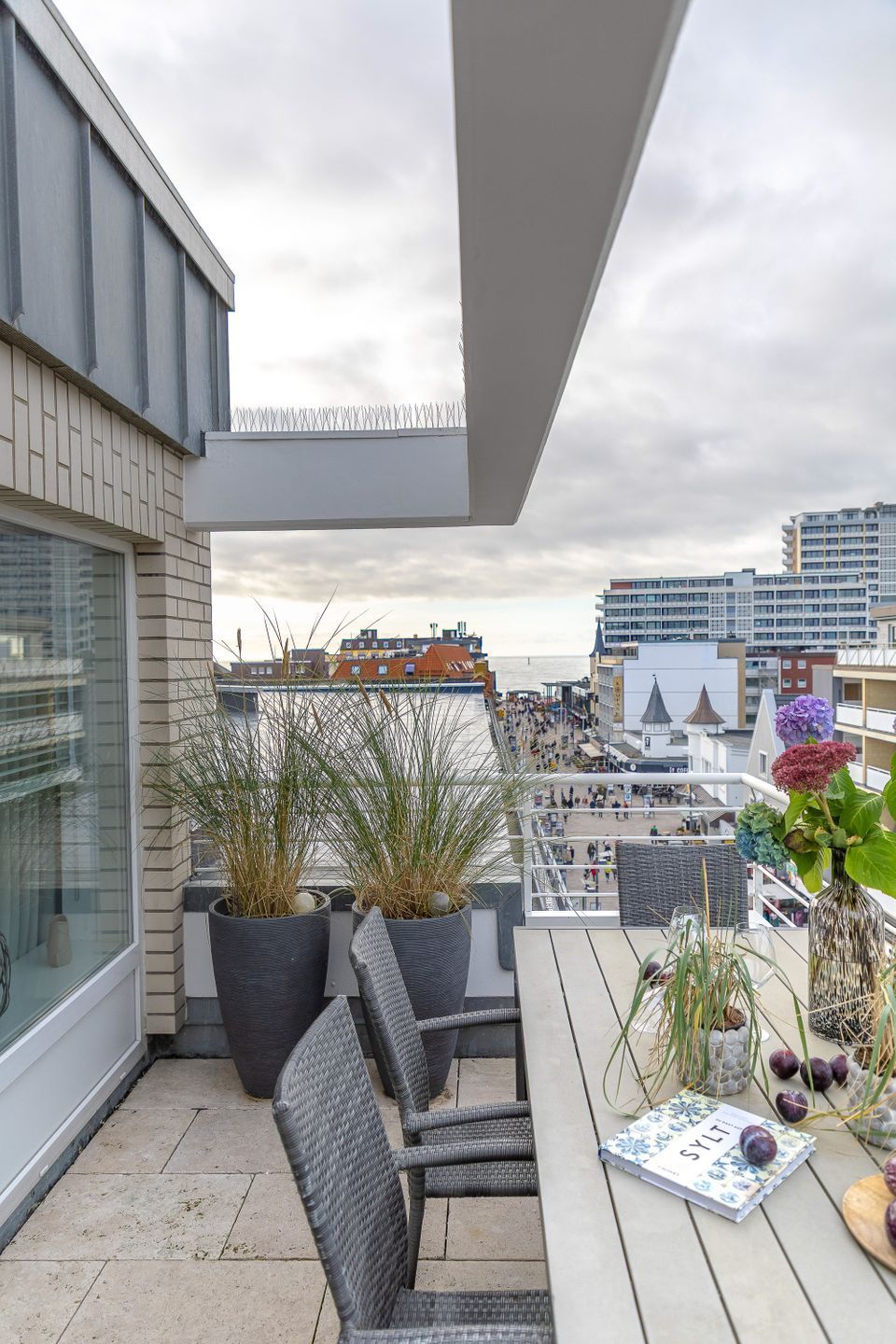A balcony with wicker chairs, a table with a book and flowers, and potted plants.