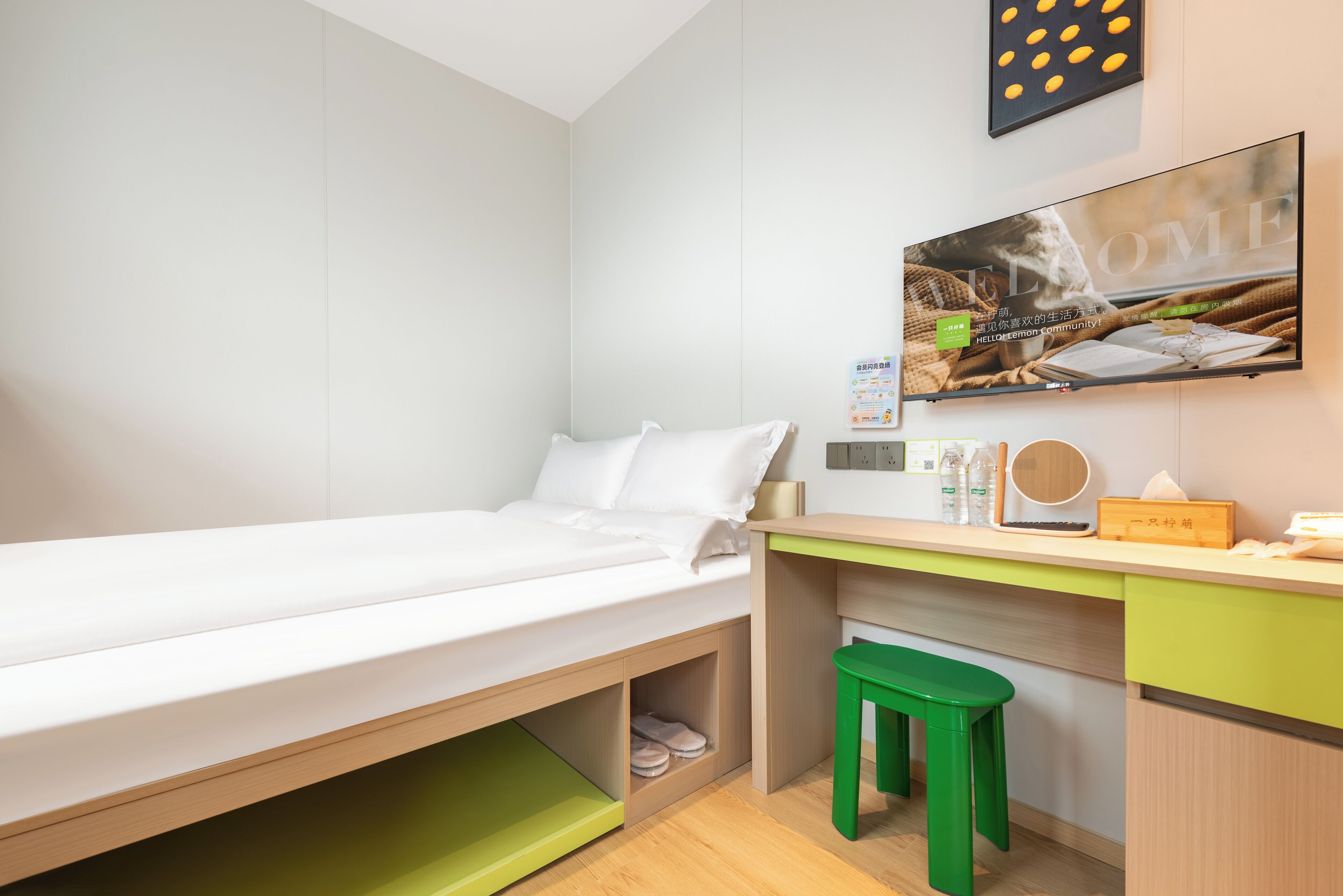 A neatly arranged bedroom with a single bed, a desk with a chair, a small stool, and a wall-mounted TV.