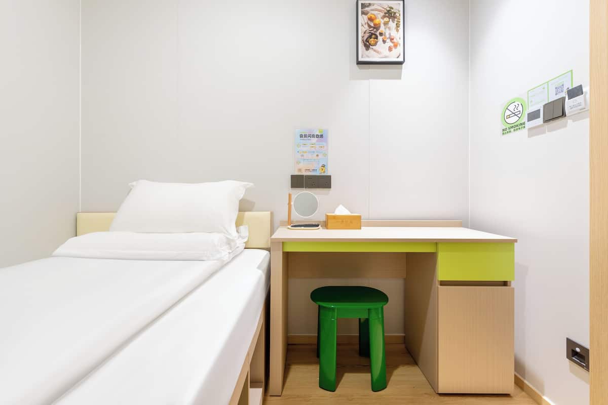 Ein kleiner, ordentlicher Raum mit einem Bett, einem Schreibtisch mit einem grünen Hocker, einer Wandladungestation und einem gerahmten Bild.