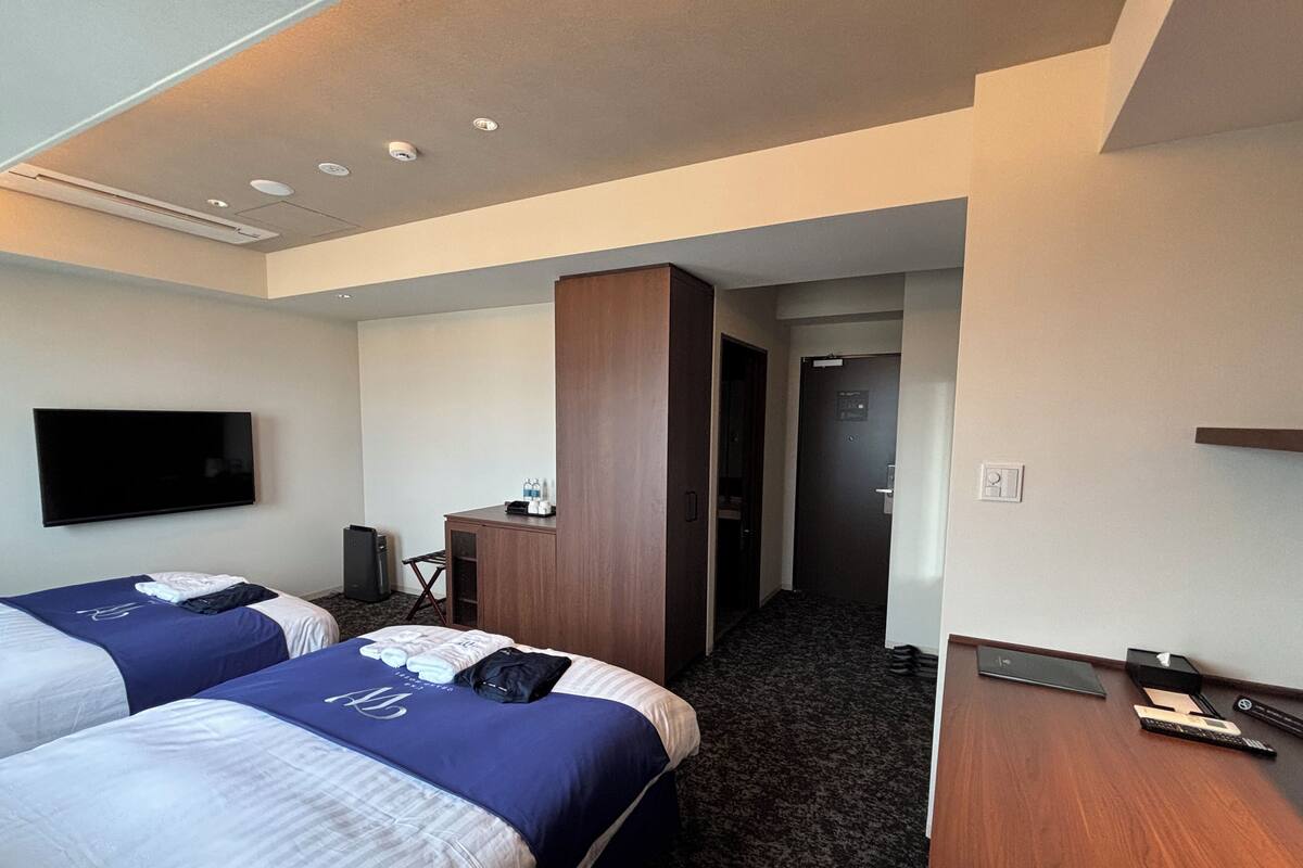 A hotel room with two beds, a flat-screen TV, a desk with a computer, and a door leading to a hallway.
