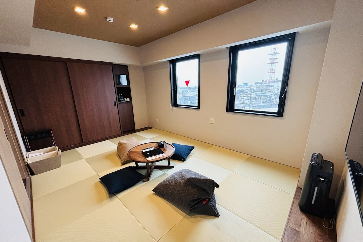 A room with tatami flooring, a low table with cushions, a wardrobe, and two windows with city views.