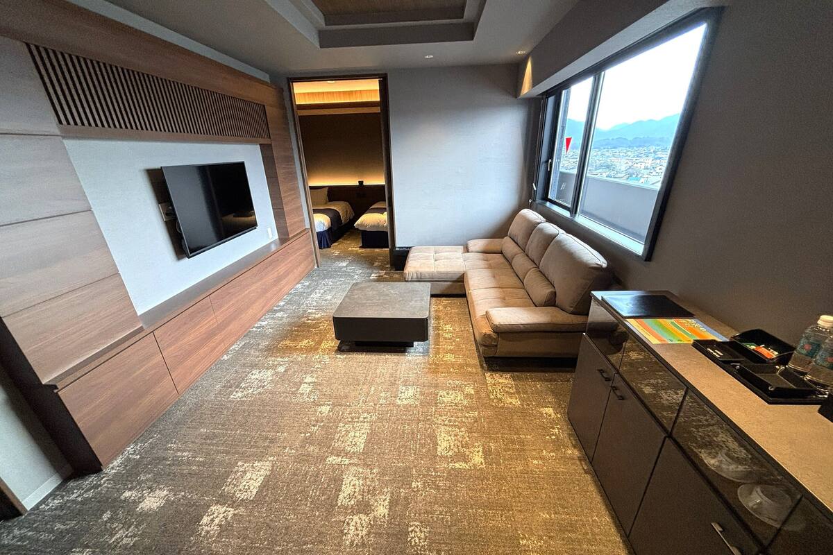 A modern hotel room with a sofa, a coffee table, a TV mounted on the wall, a window with a city view, and a minibar.