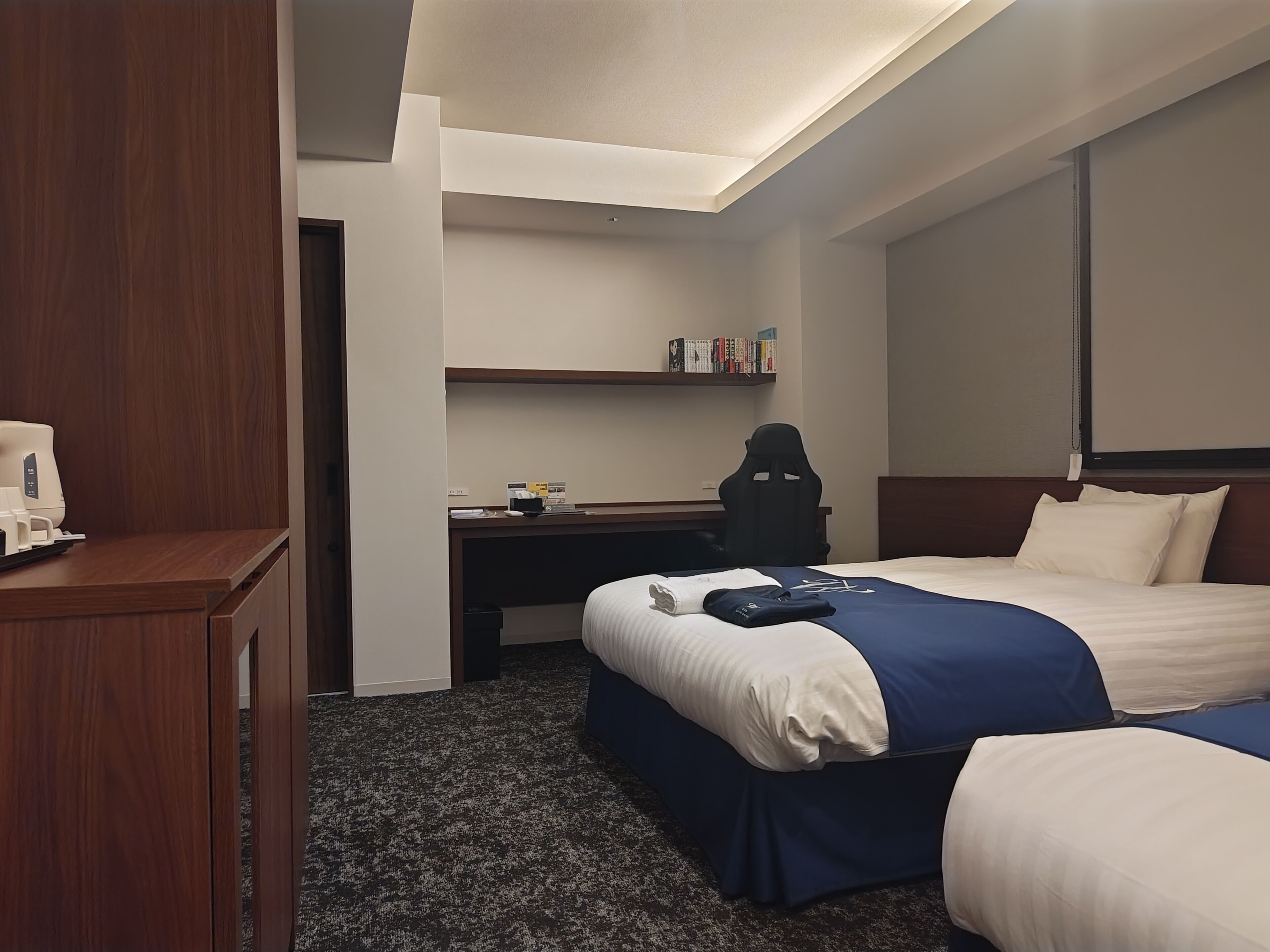 A hotel room with a desk, two beds, a bookshelf, and a coffee machine.