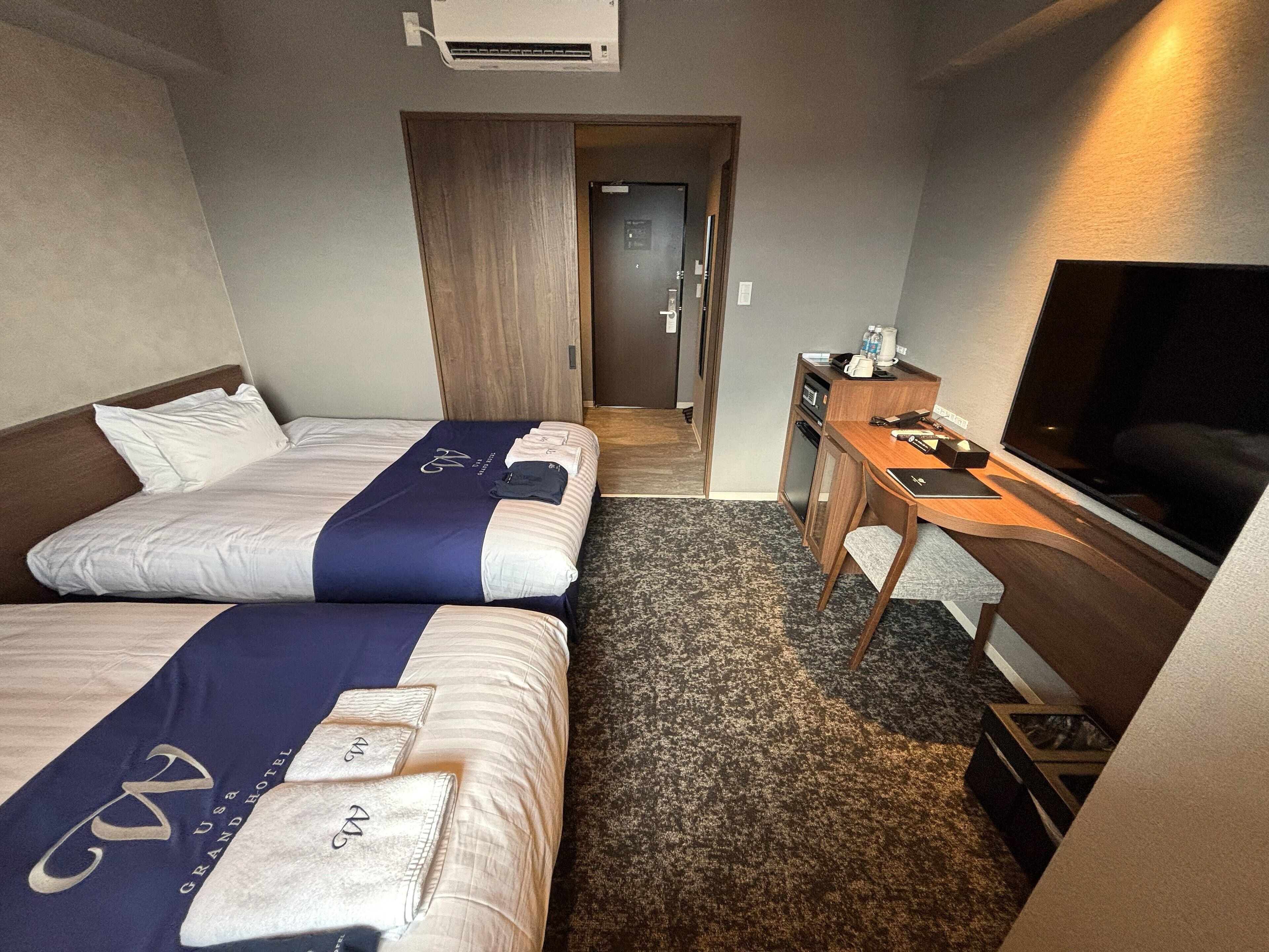 Habitación de hotel con dos camas, un escritorio, un televisor y una puerta que da a un pasillo.