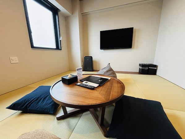 Una habitación con suelo de esteras de tatami, una mesa redonda de madera con un control remoto, una tableta y una caja de pañuelos. Hay un altavoz negro y un televisor en la pared.