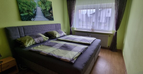 A room with two beds, a radiator, a window with curtains, and a painting on the wall.