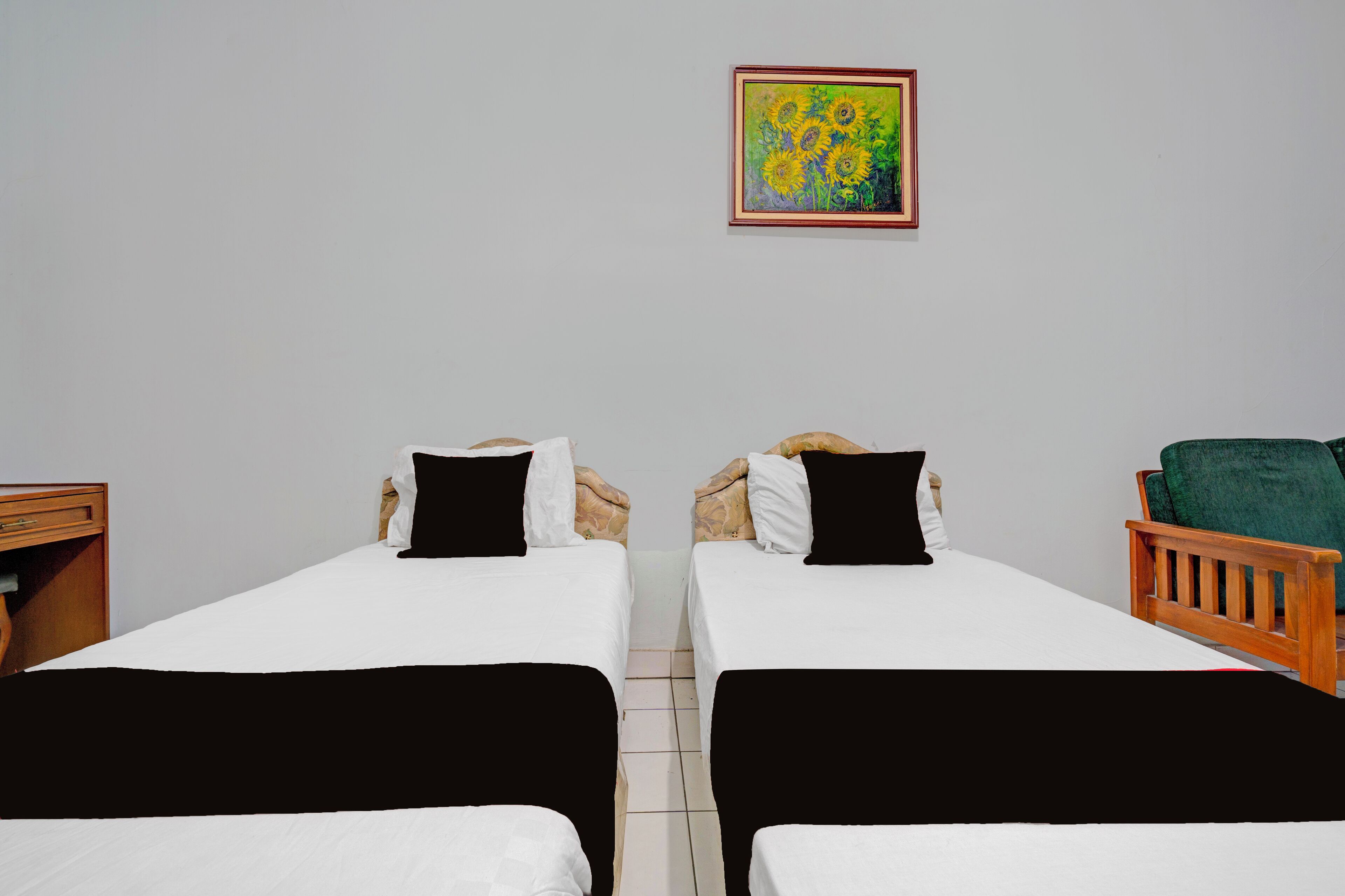 A hotel room with two single beds, a wooden nightstand, a green chair, and a painting of sunflowers on the wall.