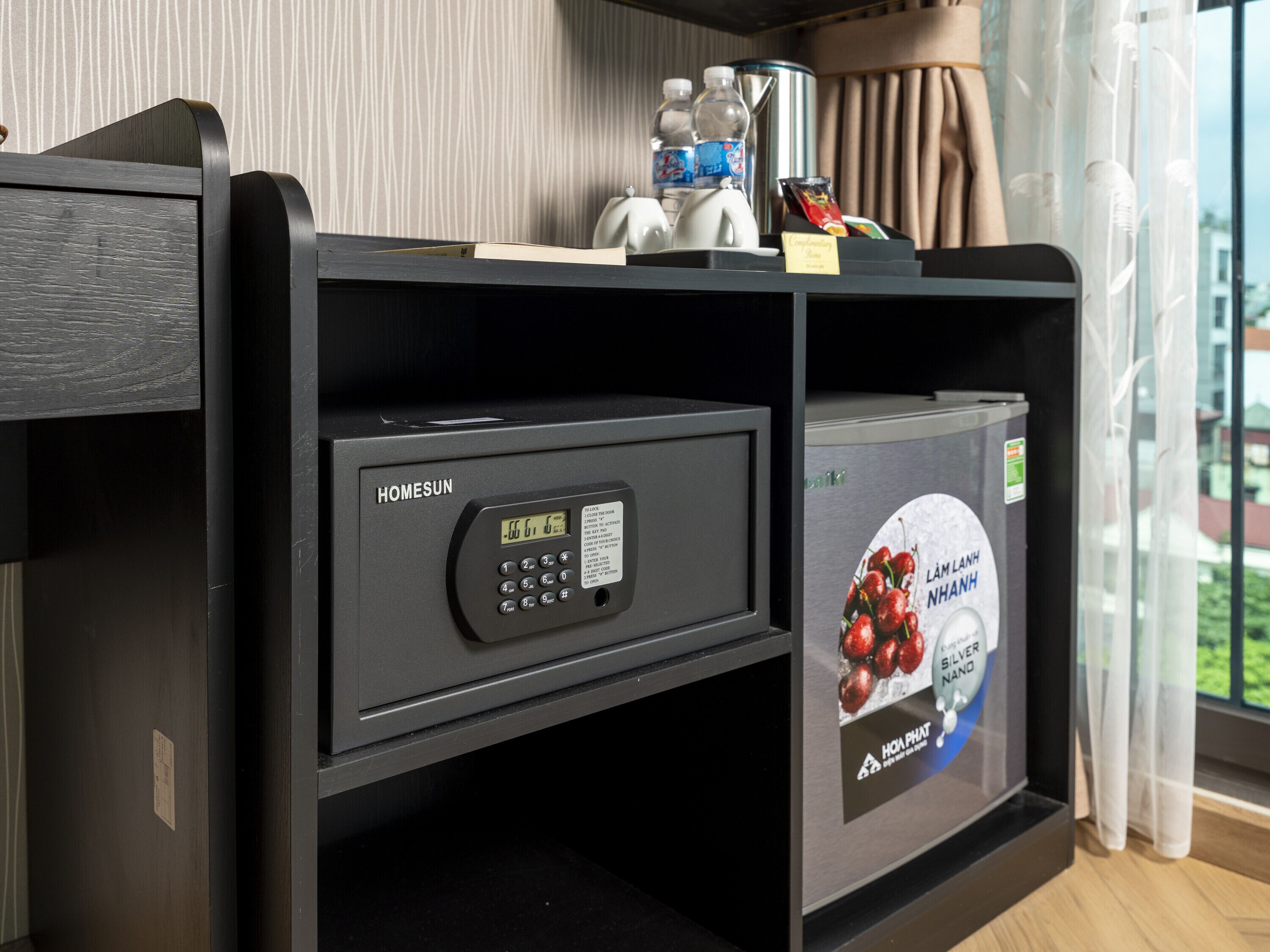 A hotel room with a minibar, a digital safe, and a refrigerator with a beverage advertisement.