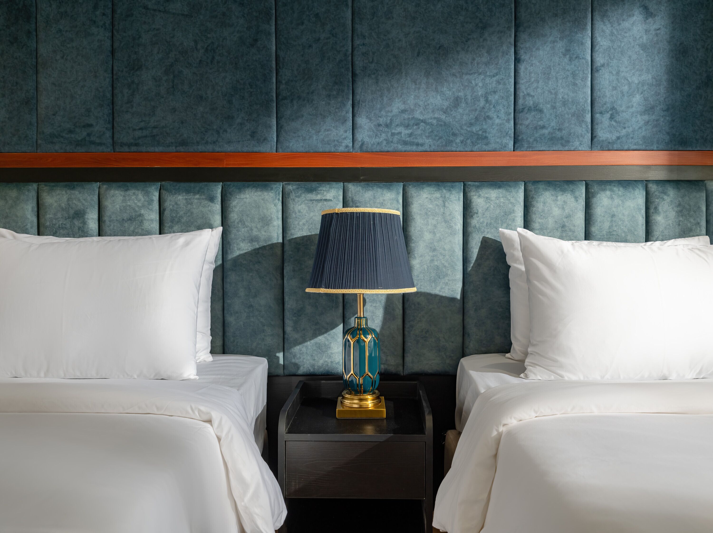 A hotel room with two beds, a dark headboard, a nightstand with a lamp, and a wooden accent strip.