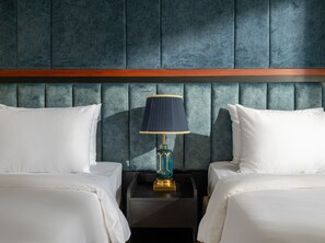 Habitación de hotel con dos camas, cabecera oscura, mesita de noche con lámpara y detalle de madera.