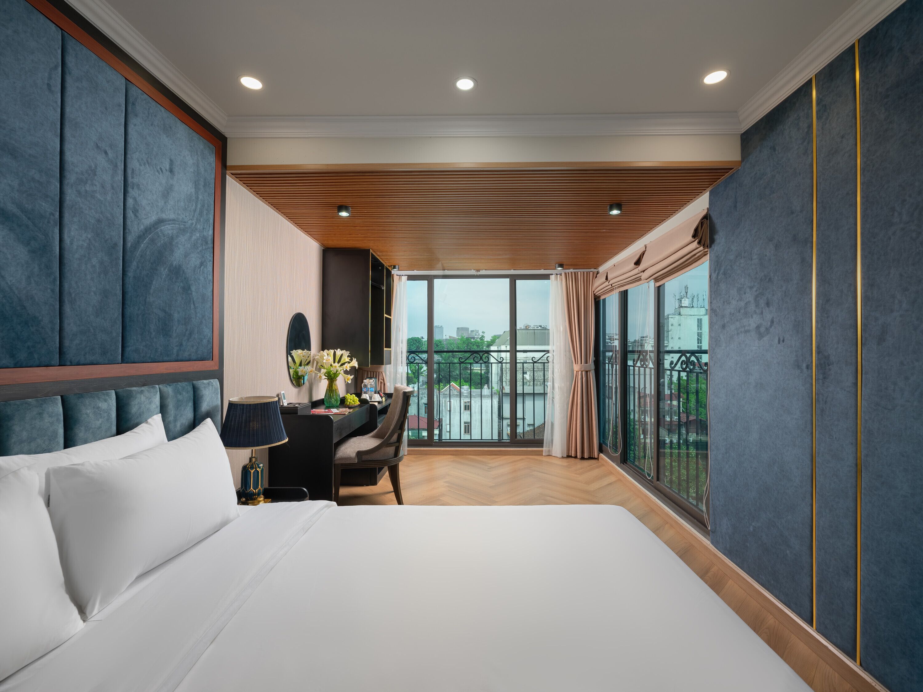 Ein Hotelzimmer mit einem großen Bett, einem Schreibtisch mit Blumenvase, Blick auf die Stadt durch große Fenster und einem Balkon mit Geländer.