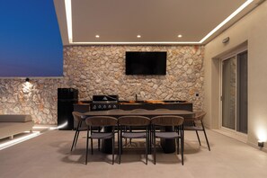 An outdoor kitchen area with a stone wall, a flat-screen TV, and a dining table with chairs.