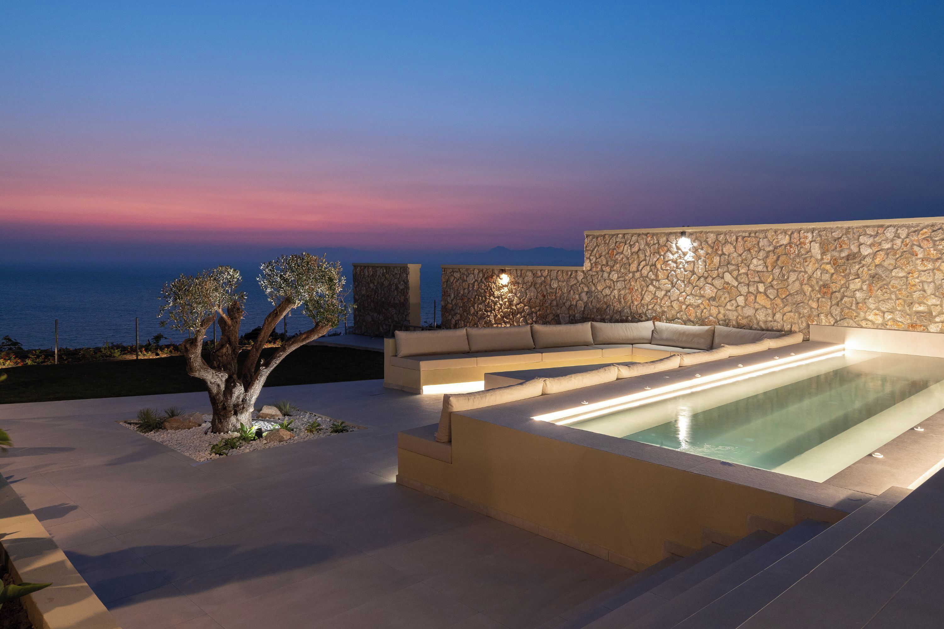 A modern outdoor pool area with a stone wall, a seating area, and a tree.