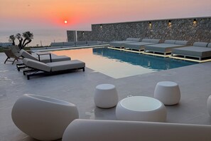 A poolside area with lounge chairs and a stone wall, overlooking the sea at sunset.