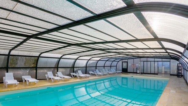 An indoor swimming pool with a curved roof structure and lounge chairs along the poolside.