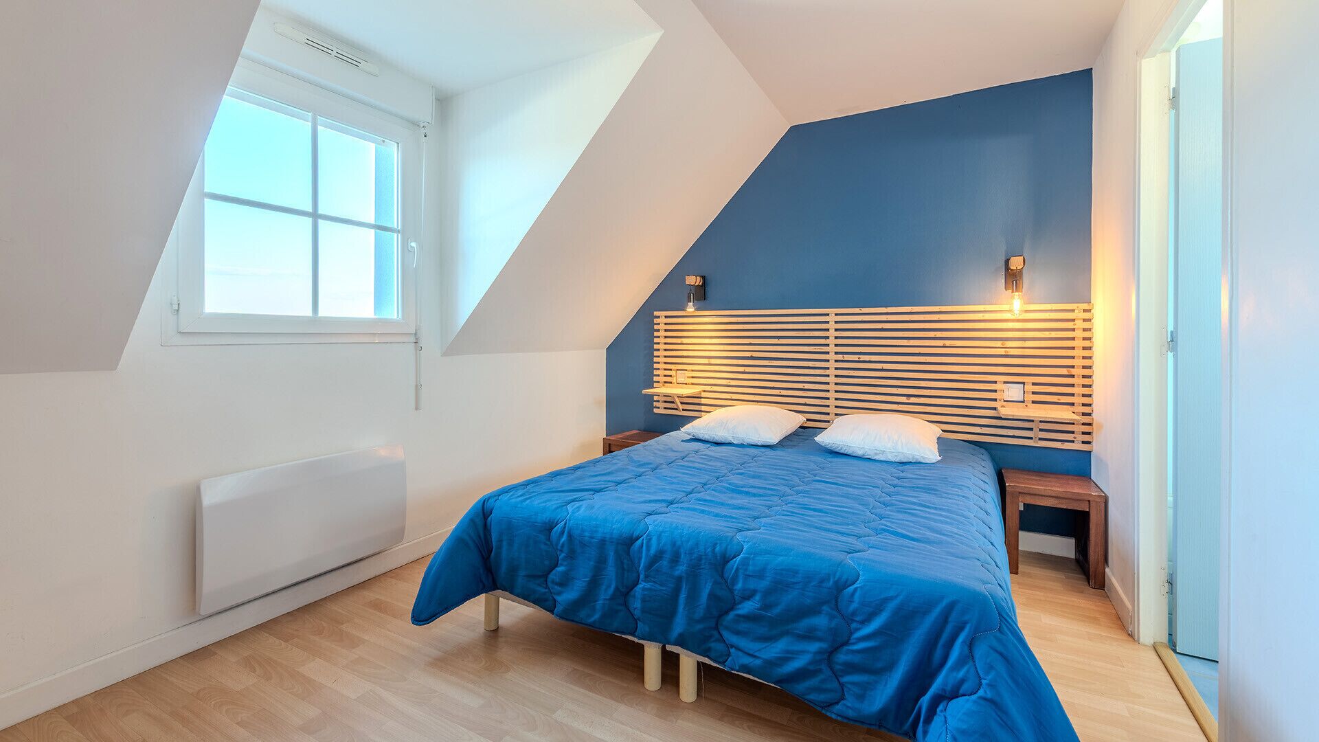 Une chambre avec un plafond en pente, un lit avec une literie bleue, deux lampes de chevet et une fenêtre.