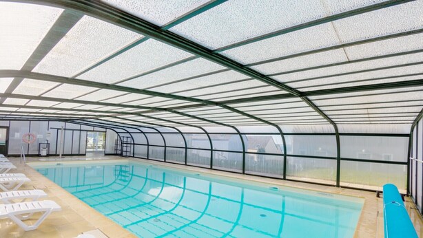 En indendørs swimmingpool med krystalklart vand, omgivet af hvide liggestole og et glasloft.