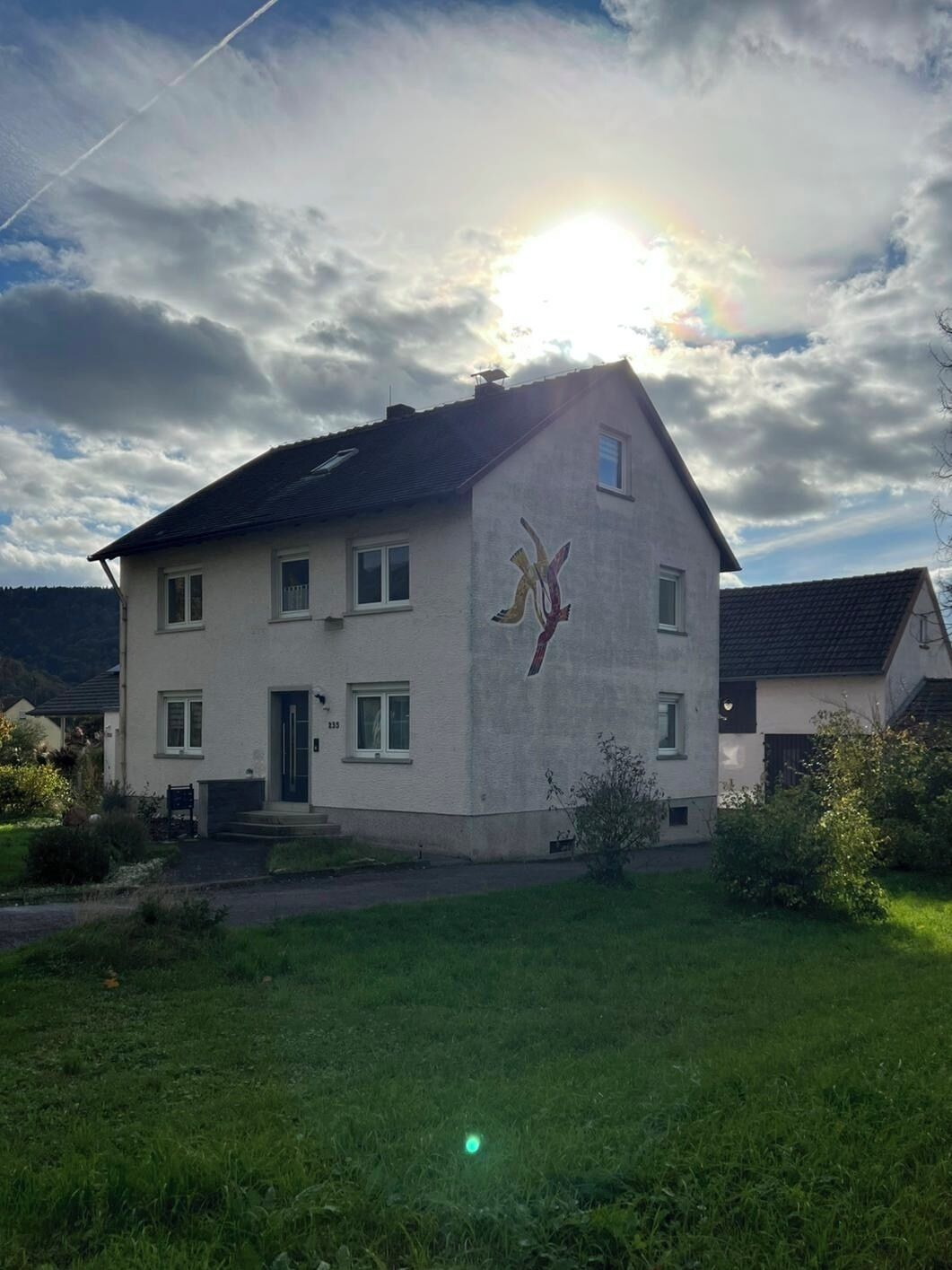A two-story house with a large mural on the side, a well-maintained lawn, and a clear sky with the sun shining.