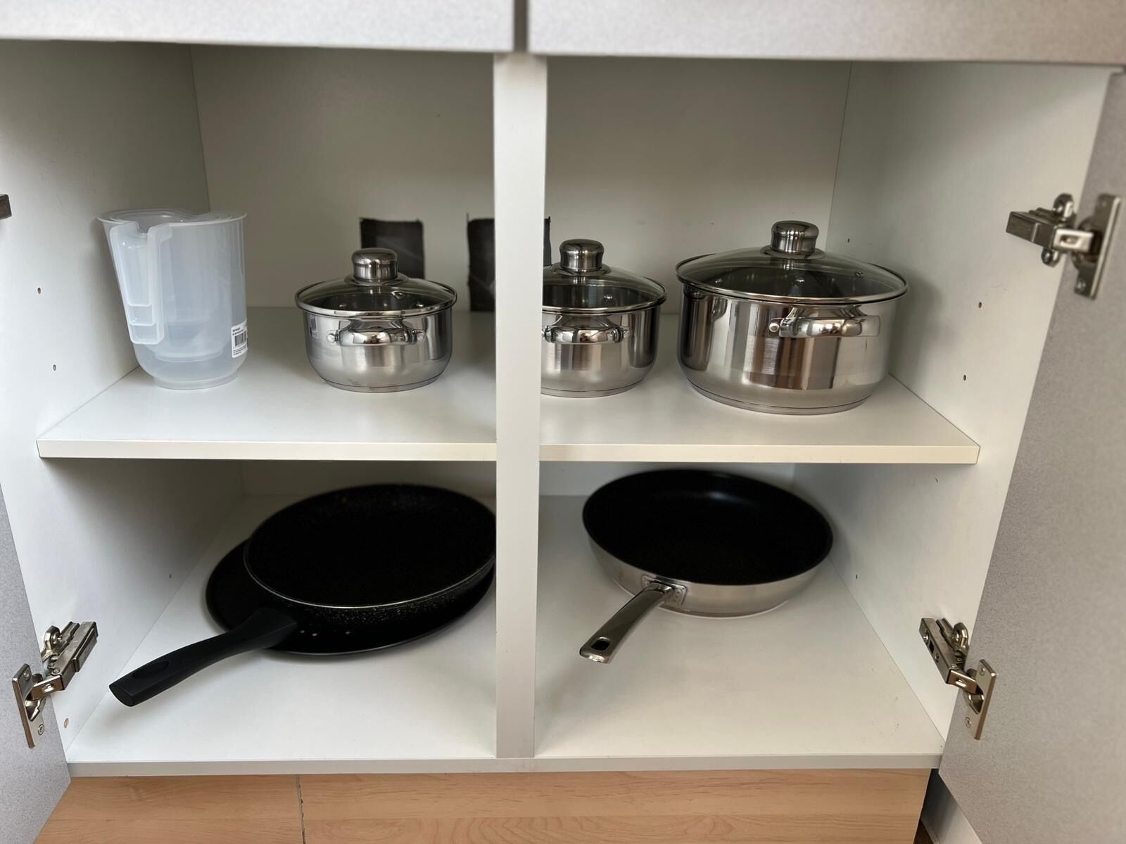Un armario de cocina con utensilios de cocina de acero inoxidable, incluyendo ollas, sartenes y una taza medidora.