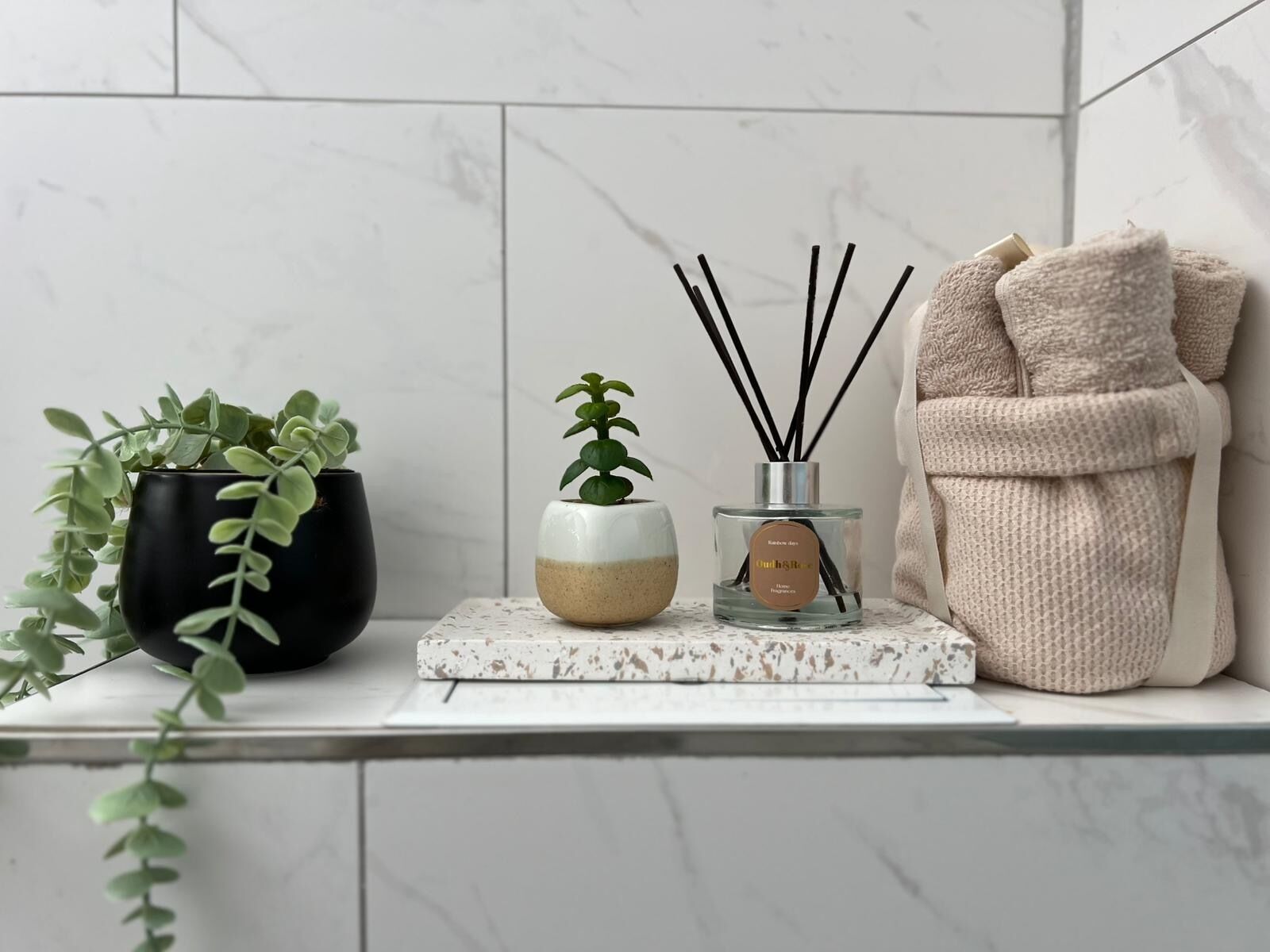 A bathroom vanity with a marble countertop, a black potted plant, a small white and beige pot with a green plant, a glass bottle with reed diffuser sticks, and a beige towel.