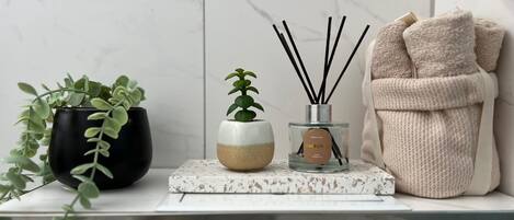 A bathroom vanity with a marble countertop, a black potted plant, a small white and beige pot with a green plant, a glass bottle with reed diffuser sticks, and a beige towel.