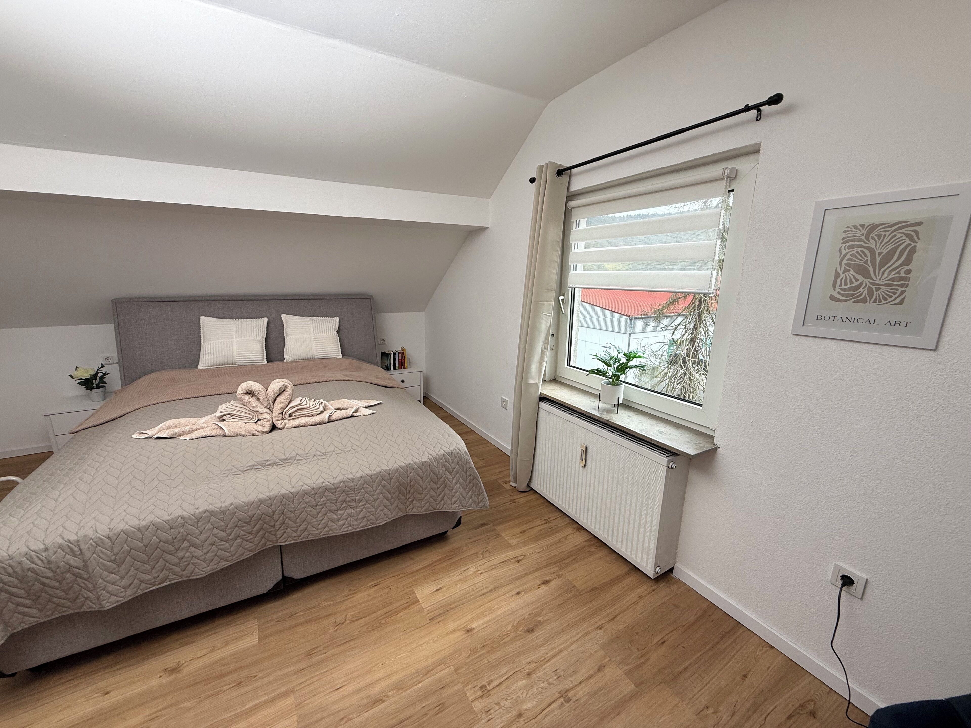 Ein Schlafzimmer mit Bett, Heizkörper, Fenster mit Blick und einem gerahmten botanischen Kunstwerk an der Wand.