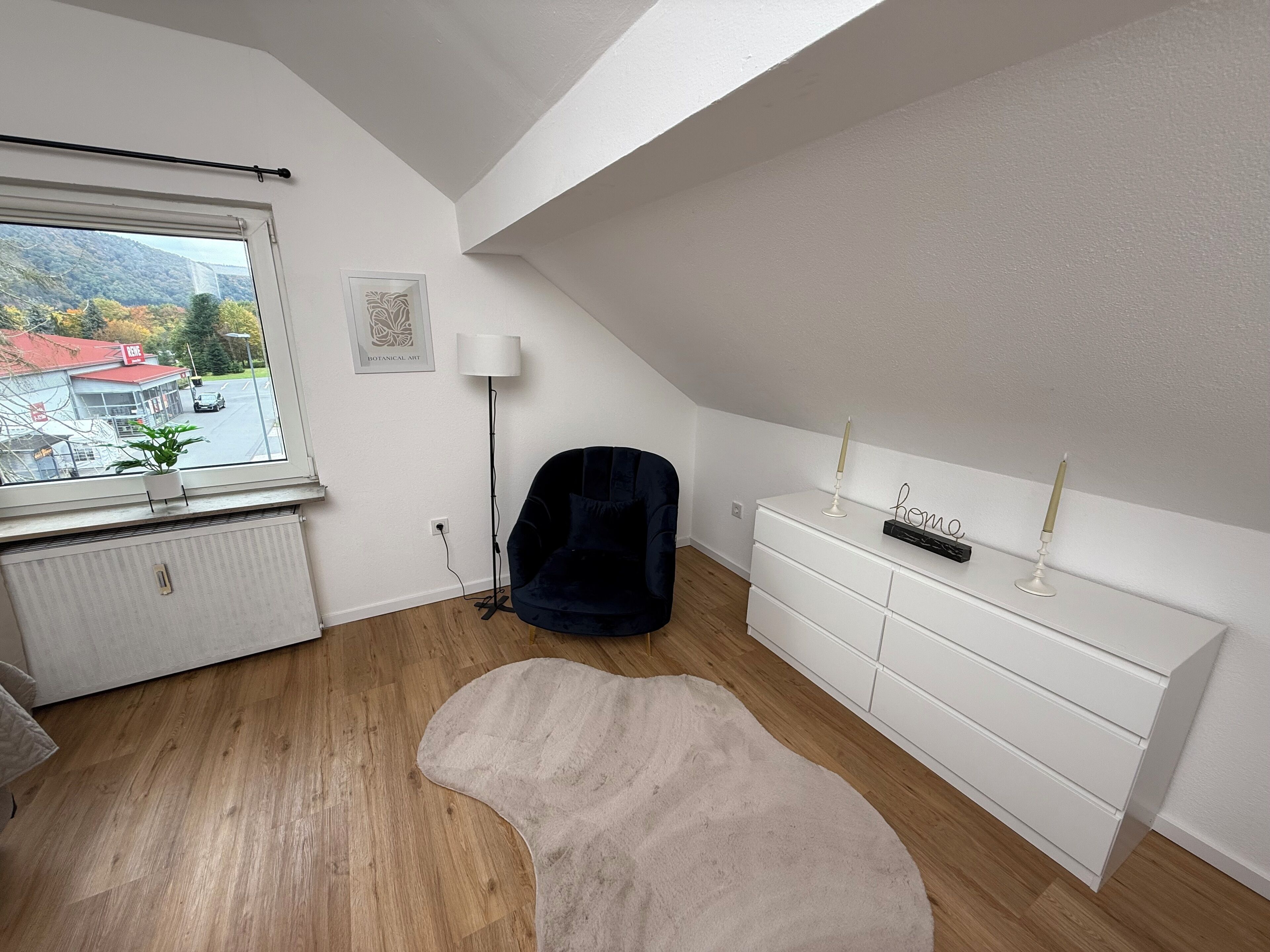 Ein modernes Zimmer mit schrägen Wänden, einem großen Fenster, einem dunkelblauen Sessel, einer weißen Kommode und einem beigen Teppich.