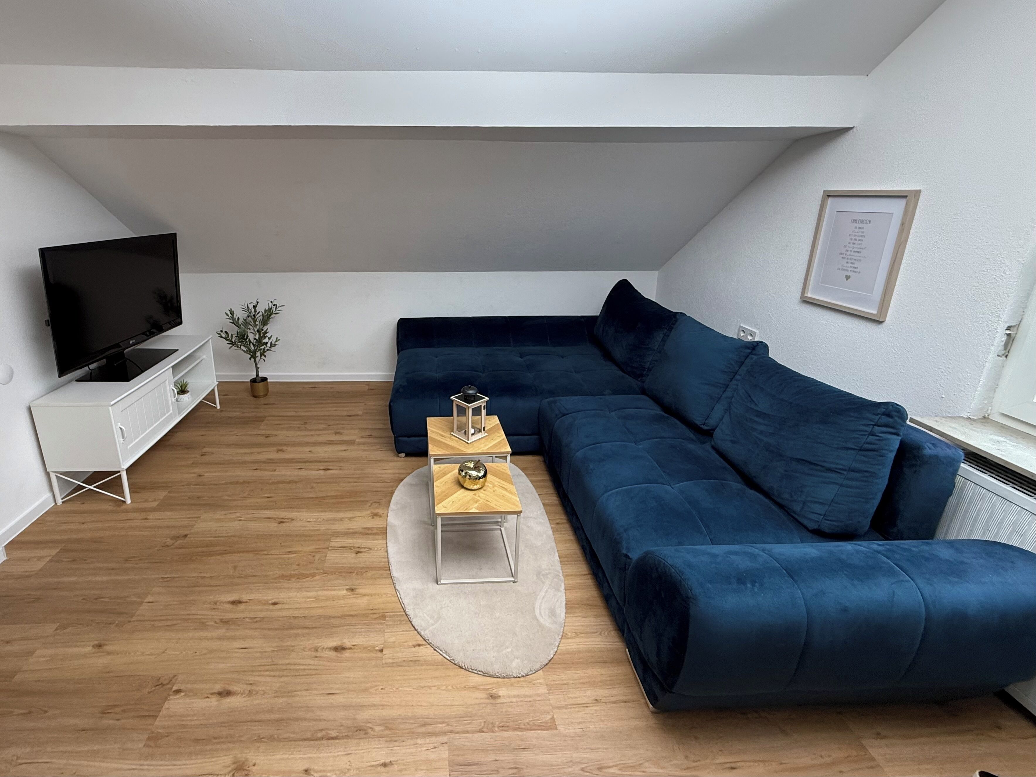 A modern living room with a dark blue sectional sofa, a white TV stand, a small wooden side table, and a potted plant.