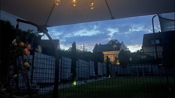 Un espace patio extérieur doté d’une structure au plafond avec des guirlandes lumineuses, offrant une vue sur le ciel et donnant sur un jardin clos.