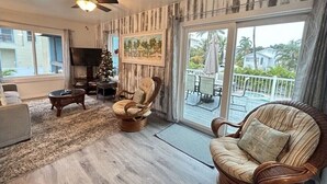 A living room with a ceiling fan, a fireplace, a television, and a large window overlooking a deck. - AMI Cortez Beach Retreat "Views" Resort-style Pool + WiFi + Dog-friendly (Bradenton Beach)