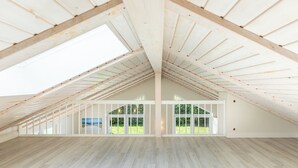 A spacious attic room with wooden beams, large windows, and a view of greenery outside.