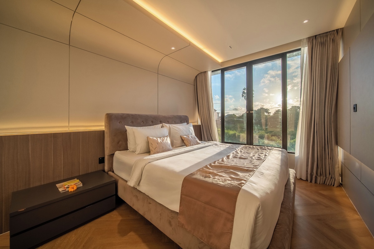 A modern bedroom with a large bed, a nightstand, and a view of the outdoors through a large window.