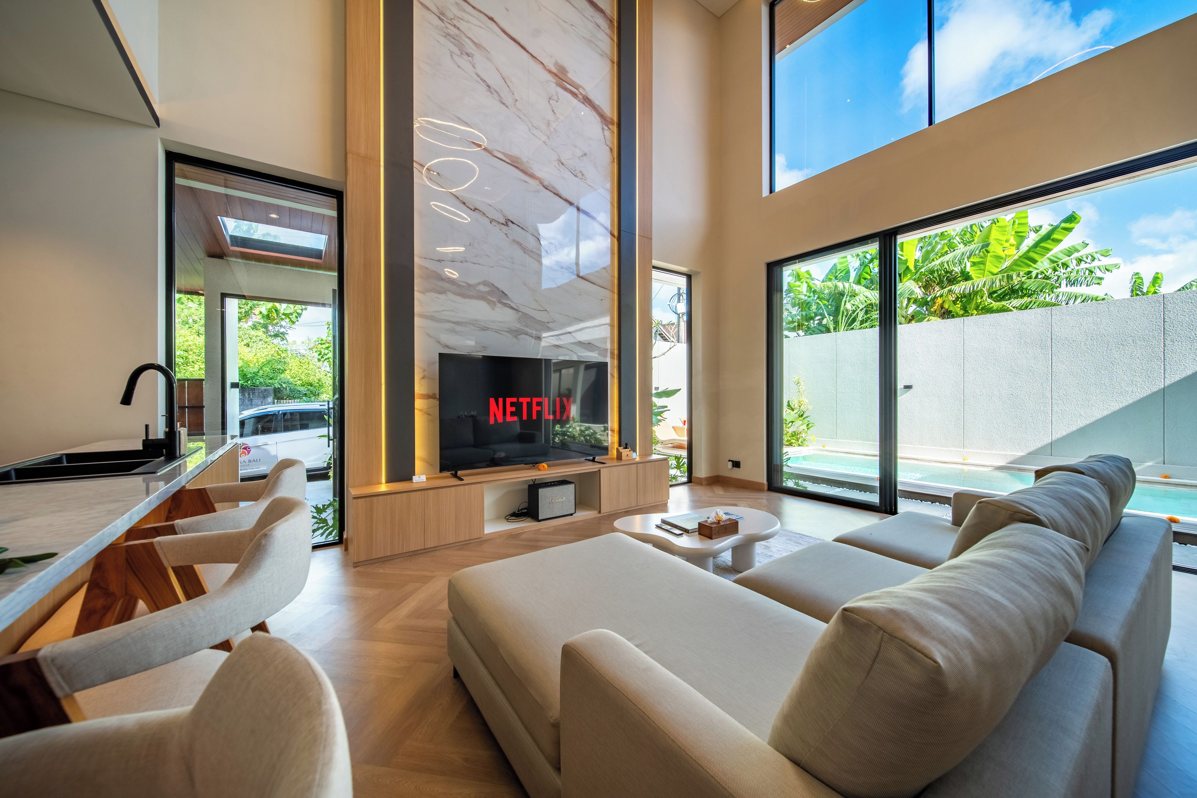 A modern living room with a large TV screen displaying 'NETFLIX,' a sofa, a coffee table, and a view of a swimming pool.